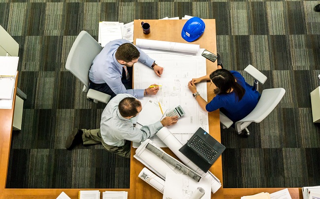 Three workers at desk, with hardhat, large maps and calculator